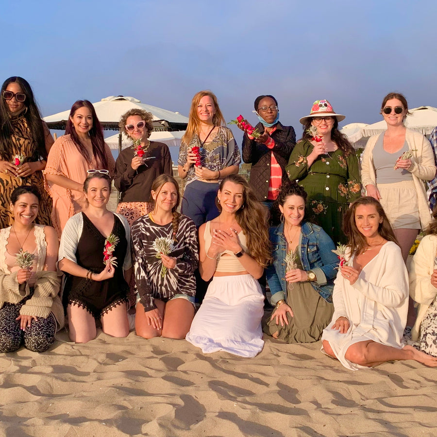 Group of smiling women holding flowers on a beach at dusk.